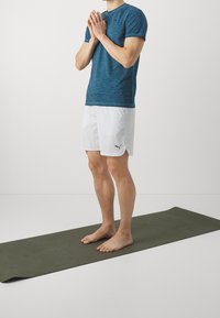 Persona descalza sobre una esterilla de yoga, vestida con una camiseta azul y pantalones cortos blancos de Puma, de pie con las manos unidas cerca del pecho.