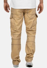 Pantalons cargo beige avec des coutures visibles, des poches latérales et une coupe ample. Le tissu semble lisse avec une finition légèrement texturée.