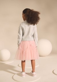 Sivý sveter a ružová tylová sukňa s dekoračnými mašľami, doplnené bielymi teniskami. Scéna obsahuje biele geometrické tvary na pozadí.