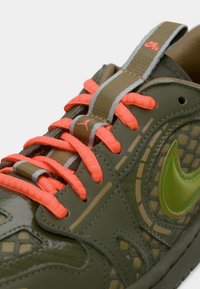 Κοντινή λήψη από ελιά-πράσινα αθλητικά παπούτσια με φωτεινά πορτοκαλί κορδόνια, ανακλαστικές γκρι λεπτομέρειες και πράσινο σήμα Nike σε υφασμάτινη επιφάνεια.