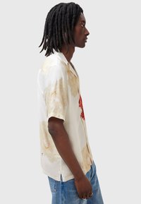 Camisa de manga corta en seda blanca con estampado floral en beige y rojo. Presenta un cuello clásico y aberturas laterales para mayor comodidad.