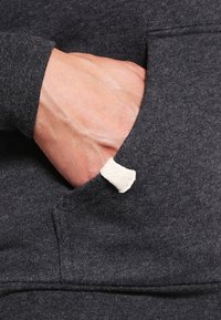 Tmavě šedý materiál mikiny s přední kapsou a látkovou záložkou. Ruka je opřená uvnitř kapsy, což ukazuje detail textury.