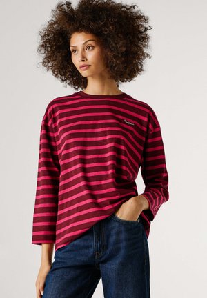 Red and pink striped long-sleeve t-shirt with a round neckline and a small logo on the left chest, paired with dark blue jeans.