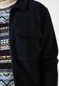 Close-up van een persoon die een gebreide trui met patroon onder een donkerblauwe wollen jas draagt, met een grote klepzak en een ritssluiting aan de voorkant.