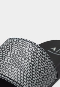 Μαύρη παντόφλα-σλάιντ με επαναλαμβανόμενο λευκό σχέδιο «AX» στο λουράκι και απλή μαύρη σόλα με το λογότυπο «AX» τυπωμένο στη σόλα του ποδιού.