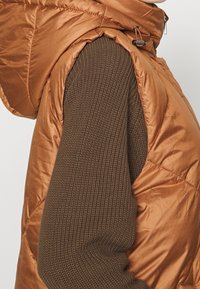 Ruskea tikkitaskuliivi, jossa kiiltävässä, tikatussa kankaassa korkea kaulus ja kiristysnyöri. Alla näkyy teksturoitu, tummanruskea neulottu neulepaita.