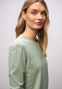 Haut à manches longues vert menthe avec un col rond et des épaules bouffantes. Le tissu semble doux et lisse, avec des détails de design minimalistes.