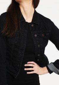 Svart denimjacka med en åtsittande design, knappstängning, bröstfickor och grå tygdetaljer på ärmarna. Texturerad tygfinish.