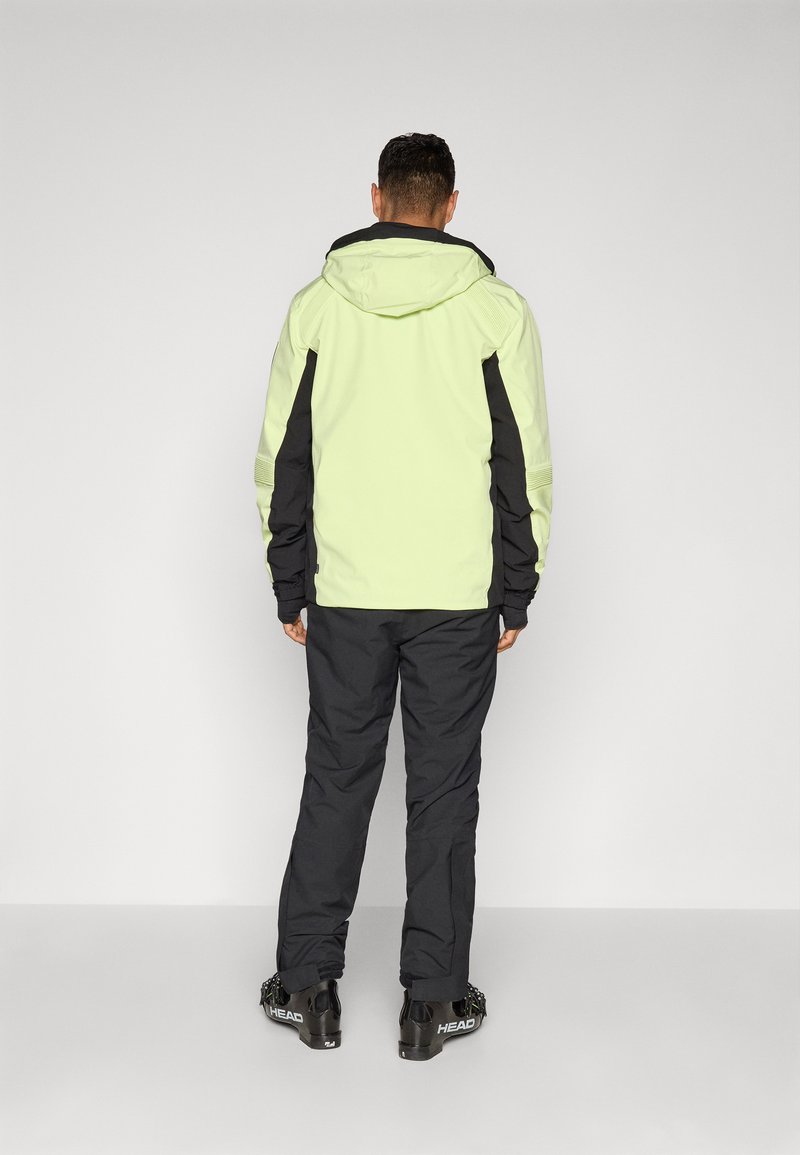 Man met de rug naar de camera, gekleed in een lichtgroene en zwarte hoodie, zwarte broek en zwarte skiboots met "HEAD"-merk.
