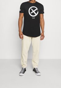 Czarna koszulka polo z krótkim rękawem z dużą białą okrągłą grafiką i tekstem z przodu; w zestawieniu z kremowymi spodniami dresowymi i czarnymi sneakersami.