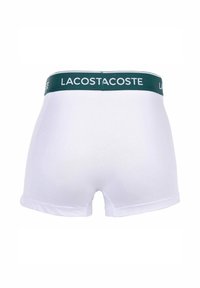 Boxer da uomo bianchi con una vita verde su cui è scritto "LACOSTE". Il tessuto appare liscio ed elastico, con un design classico e aderente.
