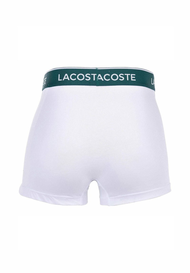 Boxers blancs avec une ceinture verte affichant "LACOSTE". Le tissu semble lisse et extensible, avec un design classique et ajusté.