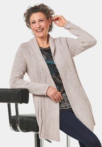 Hellrosa, gestrickte Strickjacke mit geripptem Muster und offenem Frontdesign. Getragen über einem gemusterten Oberteil, das einen lässigen Schnitt und lange Ärmeln hat.