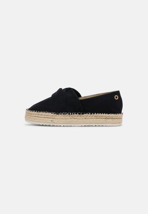 Sort espadrille slip-on med flettet juttersål og lille metaløje nær hælen, vist i sideprofil på hvid baggrund.
