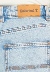 Shorts en denim avec un délavage bleu clair, ornés de surpiqûres orange et d'une étiquette en cuir tan Timberland sur la ceinture. Poche arrière visible.