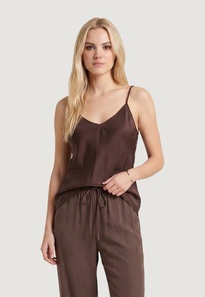 Giovane donna con lunghi capelli biondi che indossa un top marrone senza maniche con spalline sottili e pantaloni marroni abbinati su uno sfondo neutro.