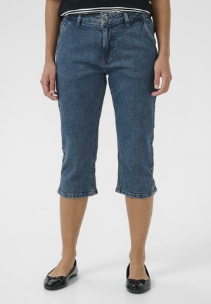 Osoba ubrana w niebieskie dżinsowe spodenki capri o średnim stanie z podwiniętymi nogawkami i czarne baleriny na białym tle.