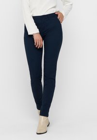 Námořnické modré slim-fit kalhoty s plochým předním dílem, poutky na opasek a bočními kapsami, v kombinaci s bílým dlouhým rukávem a světlými botami.