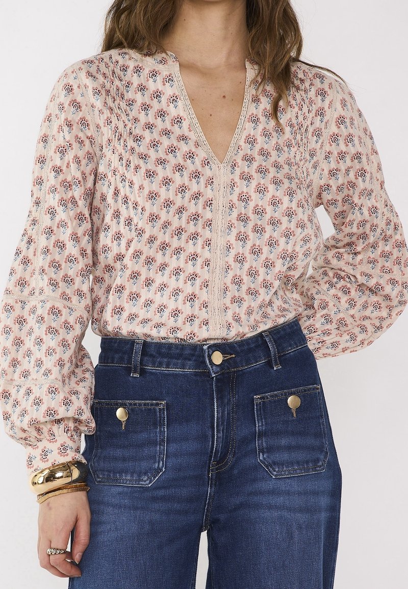 Femme portant un chemisier crème à manches longues avec un petit motif floral et un jean bleu taille haute avec poches plaquées à l'avant et boutons dorés.