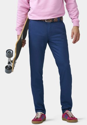 Personne portant un pantalon bleu, un sweat-shirt rose, une ceinture à motifs et des baskets rouges tenant un skateboard à côté d'elle devant un fond uni.