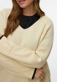 Femme portant un pull crème en V sur un haut noir, avec de longs cheveux bruns et une bague en argent à la main gauche.