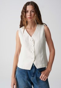 Ärmelloses weißes Top mit V-Ausschnitt, fünf Knöpfen und taillierter Silhouette. Kombiniert mit blauen Jeanshosen mit Seitentaschen.