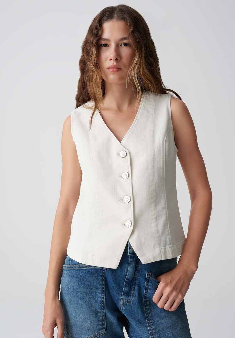 Ärmelloses weißes Top mit V-Ausschnitt, fünf Knöpfen und taillierter Silhouette. Kombiniert mit blauen Jeanshosen mit Seitentaschen.