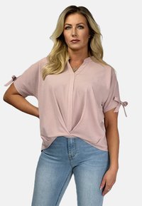 Jasnoróżowa bluzka z krótkim rękawem, dekoltem v i szczegółem w postaci skręconego przodu, z wiązanymi rękawami oraz miękką, gładką teksturą. W połączeniu z jasnoniebieskimi dżinsami.