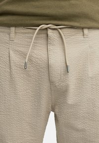 Beiga texturerade byxor med dragsko i midjan. Tyget har ett rutmönster och metallaccenter i ändarna av dragskon.