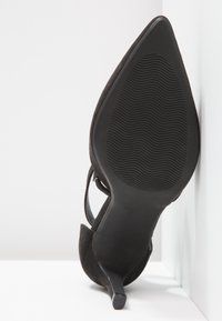 Chaussure à talon haut en suede noir avec un bout pointu, un fin talon, et une semelle texturée ondulée. Détail de la sangle de cheville réglable visible.