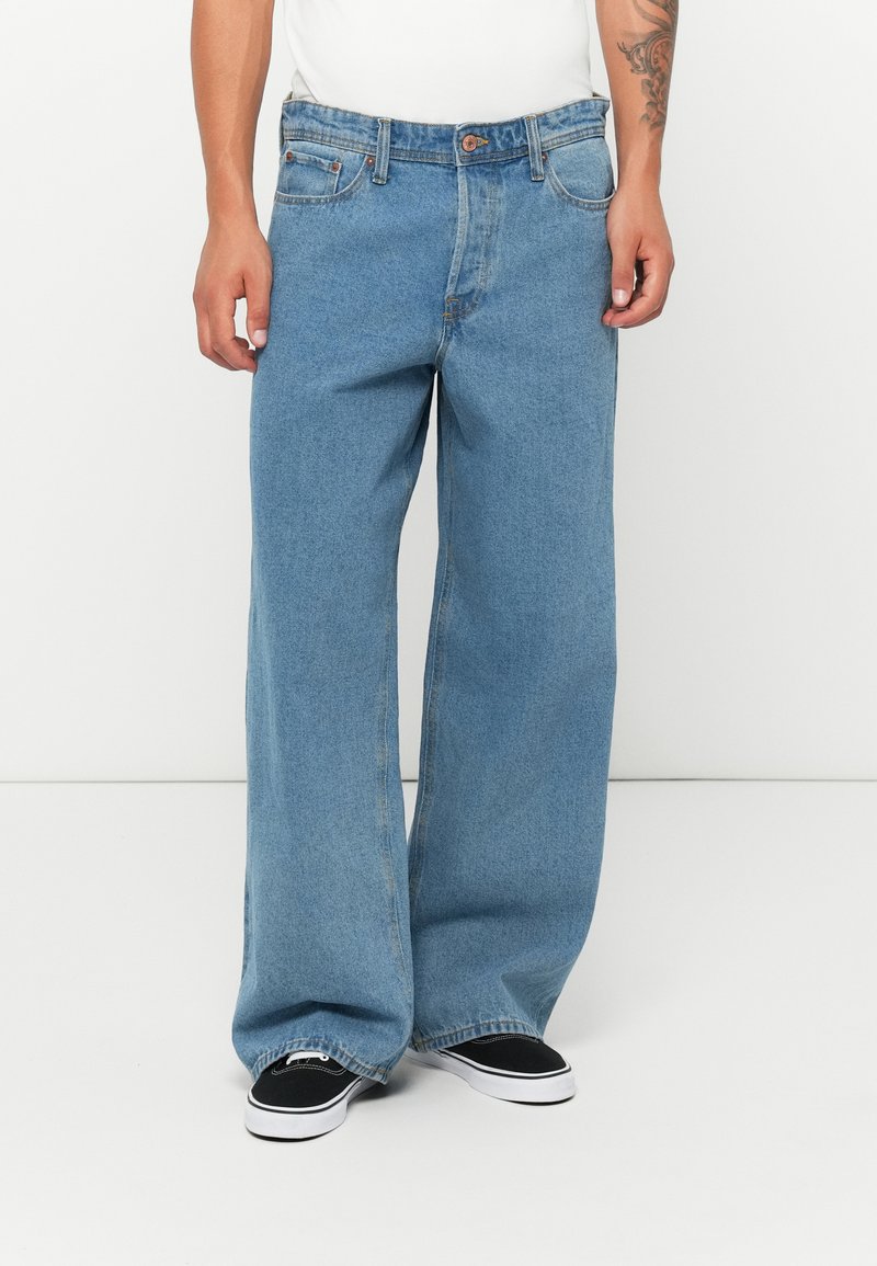 Mann i lyseblå, vide jeans og svarte sneakers står mot en hvit vegg.