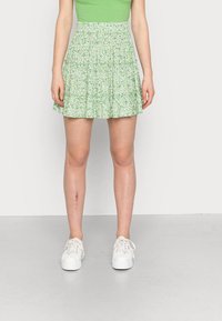 Vihreä, kerroksellinen minihame, jossa on kukkakuosi, jossa on valkoisia päivänkakkaroita. Yhdistettynä valkoisiin lenkkareihin neutraalilla taustalla.