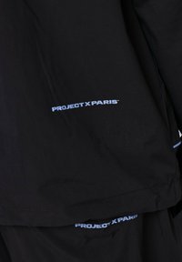 Chaqueta negra con el logo "PROJECT X PARIS" en azul claro en el lado izquierdo. Textura suave, corte moderno, diseño minimalista.