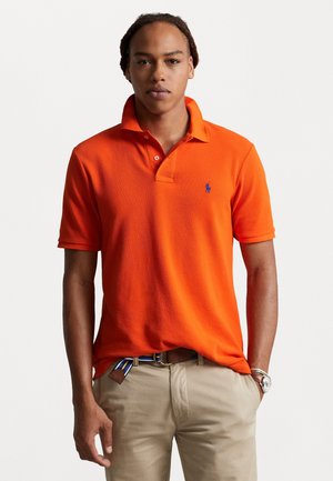 Oranje poloshirt met een klassiek ontwerp, korte mouwen, een kraag en een klein blauw logo op de linker borst, gecombineerd met beige broek.