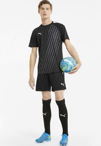Camiseta deportiva negra con rayas diagonales grises, pantalones cortos negros, calcetas negras hasta la rodilla y tacos azules. El jugador sostiene un balón de fútbol multicolor.