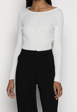 Vrouw draagt een witte geribbelde top met lange mouwen, ingestopt in een hoogtailles zwarte broek, staand tegen een effen achtergrond.