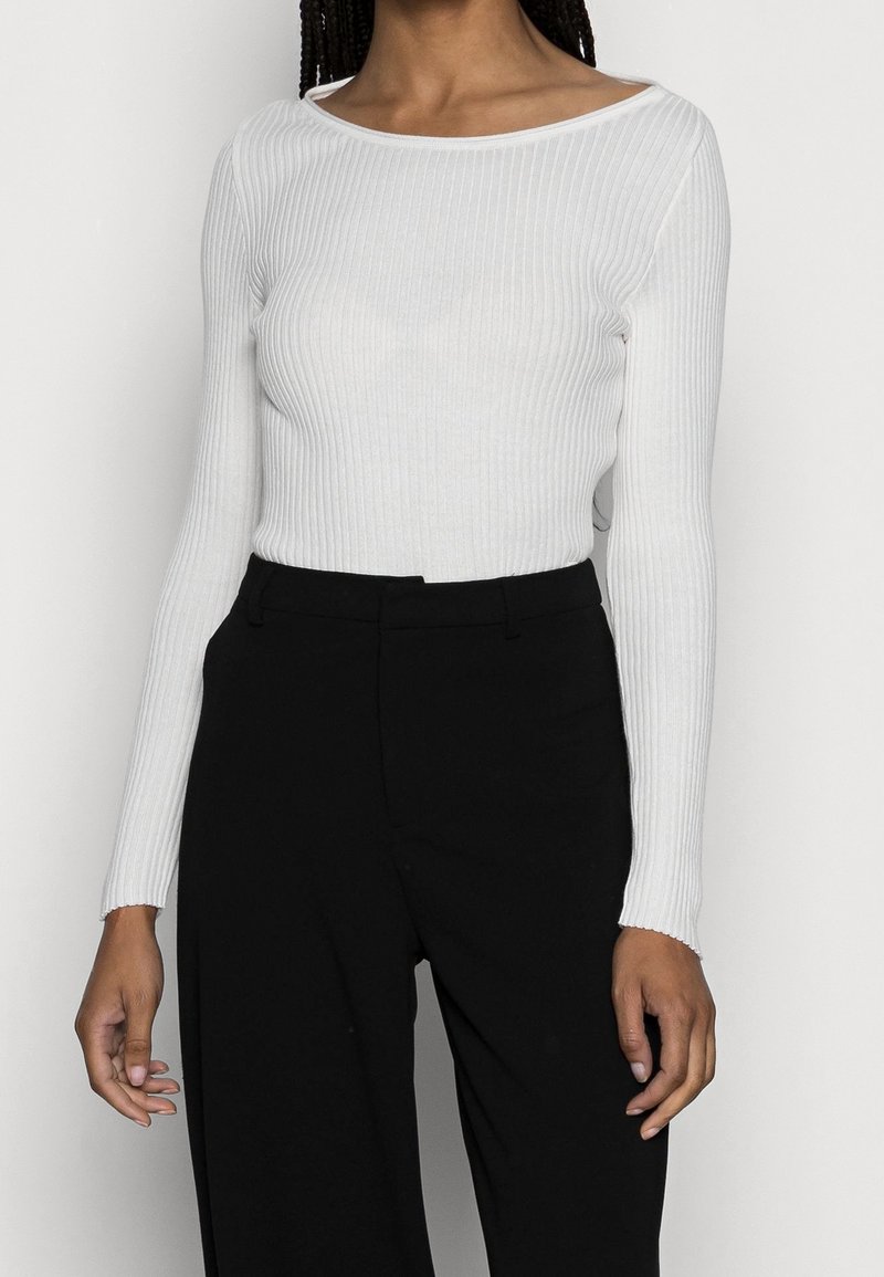 Femme portant un haut blanc côtelé à manches longues rentré dans un pantalon noir taille haute, debout devant un fond uni.