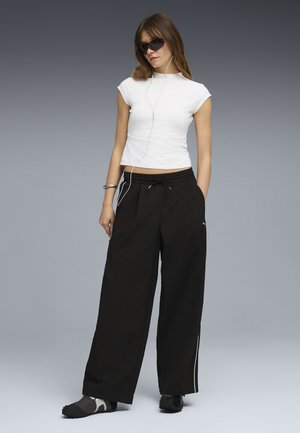 Young woman wearing black wide-leg pants, white fitted sleeveless top, black sunglasses, earphones, and black sneakers, standing against gray background.