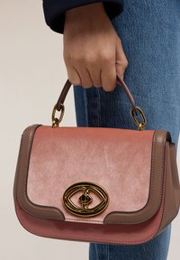 Borsa in velluto rosa strutturato e pelle marrone con chiusura ovale dorata e manico superiore, tenuta da una mano su uno sfondo di tessuto denim.