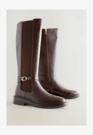 Een paar donkere bruin leren laarzen tot de knie met een zijrits, elastisch paneel en decoratieve gesp op een lichte achtergrond.