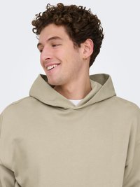 Sweat à capuche beige à coupe décontractée, montrant un col arrondi et une texture douce, porté sur un t-shirt blanc. Boucle d'oreille dorée visible.