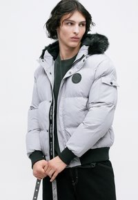 Jachetă puffer gri cu fermoar în față, glugă căptușită cu blană, manșete elastice și un patch rotund pe piept. Textură moale; design matlasat.