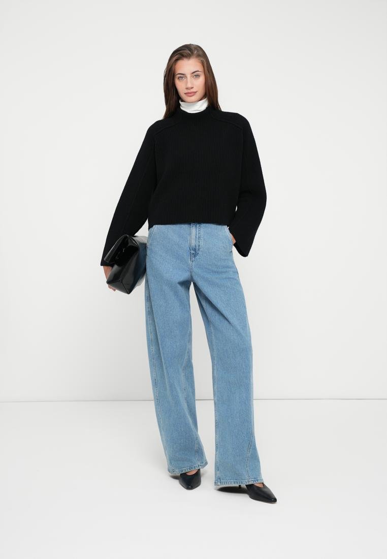 Pull noir côtelé à manches larges, associé à un jean large taille haute bleu clair. Le mannequin tient une pochette noire et porte des escarpins pointus.