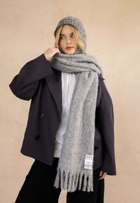 Ensemble de chapeau en laine gris et écharpe associé à un blazer foncé sur un haut blanc. L'écharpe présente des franges et une étiquette en tissu.