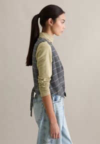 Szara kamizelka w kratę z guzikami, zestawiona z jasnooliwkowym golfem i wysokim stanem niebieskich dżinsów. Gładki materiał i dopasowany krój.