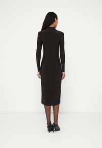 Fekete hosszú ujjú ruha, karcsúsított fazonnal, térdig érő szegéllyel és kerek nyakkivágással. Fekete magas sarkú cipővel viselve. Sima textúrájú.