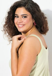 Canotta verde chiaro con spalline larghe, texture morbida. Il modello ha capelli ricci e indossa orecchini a cerchio rossi, mostrando una posa rilassata.