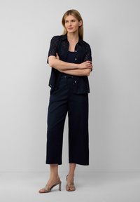 Marineblaue, durchsichtige Bluse mit Blattmuster, kombiniert mit weit geschnittenen Marineblauen Hosen. Beigefarbene Blockabsatzsandalen runden das Outfit ab.
