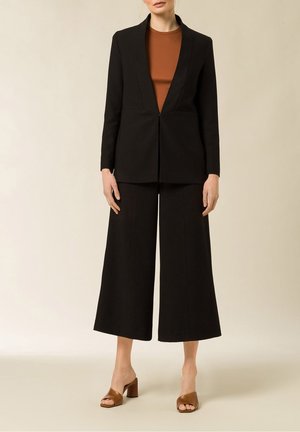 Frau trägt schwarzen, maßgeschneiderten Blazer, passende weit geschnittene, gekürzte Hose, braunes Oberteil und braune, offene Sandalen mit Absatz, steht vor hellem Hintergrund.