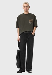 Zielona bawełniana koszulka z nadrukiem na piersi, zestawiona z czarnymi, wysokim stanem jeansami i czarną torebką. Modelka trzyma czarną torebkę.